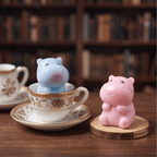 Two cute hippo-shaped ceramic tea infusers in pastel blue and pink, displayed on a wooden table with a blurred bookshelf background. One hippo is sitting inside an ornate white teacup with gold detailing, while the other sits on a round wooden coaster.