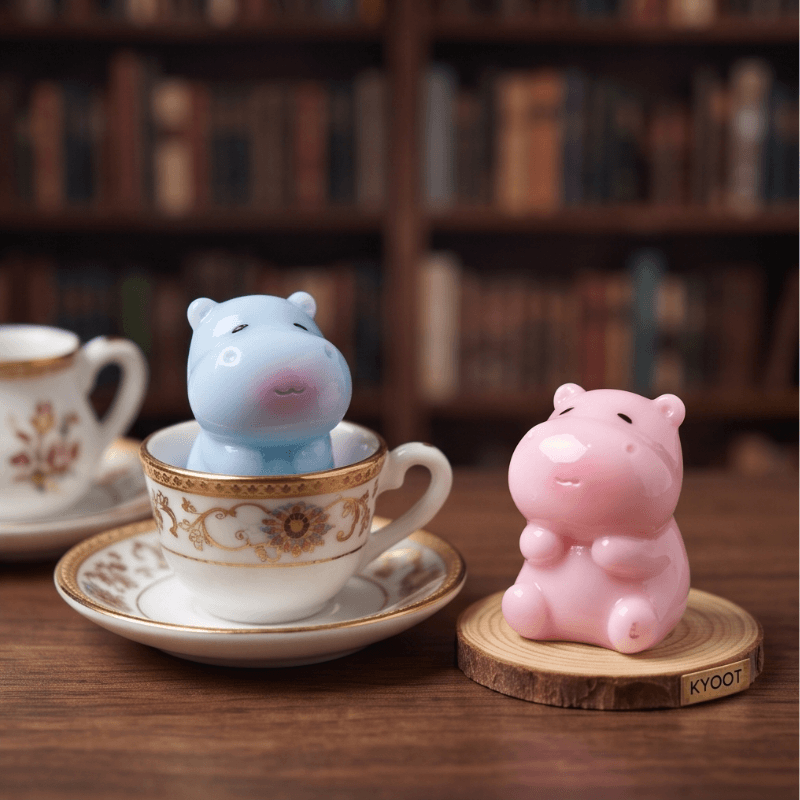 Two cute hippo-shaped ceramic tea infusers in pastel blue and pink, displayed on a wooden table with a blurred bookshelf background. One hippo is sitting inside an ornate white teacup with gold detailing, while the other sits on a round wooden coaster.