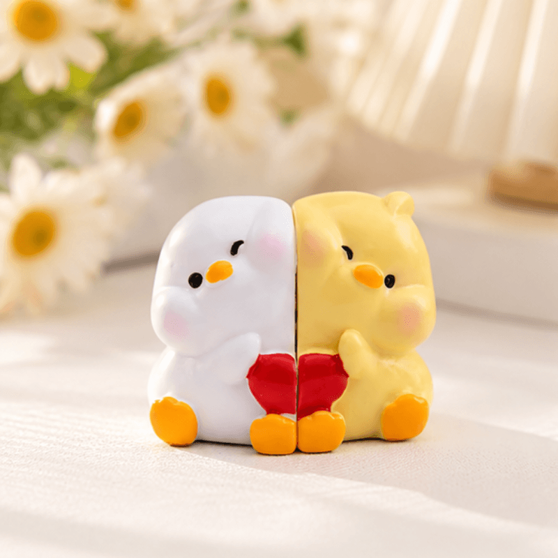 A split-color novelty object featuring a white left half with a pink cheek and yellow beak, and a yellow right half with a pink cheek and yellow beak, holding a red heart. The object is styled on a white surface with a soft-focus background of white daisies and a warm light source.