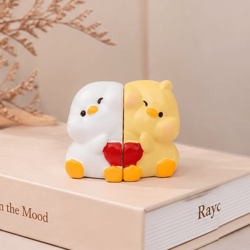 Two cute split-face duck figurines sitting side by side on a book. One half is white with a yellow beak and feet, and the other half is yellow with a white beak and feet, holding a red heart.