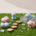 Four small hippo figurines in pastel pink and light blue are arranged on artificial green grass with clear glass stones and round wood slice props. One blue hippo is sitting in a clear shallow dish with water splashing around it.