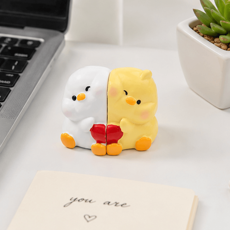 A cute split-color novelty desk accessory featuring a white duck on the left and a yellow duck on the right, holding a red heart. The item is styled on a white desk with a laptop, a succulent, and a card that reads 'you are'.
