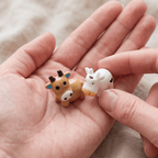 A tan cow figurine and a white spotted cow figurine held in a hand, showing their size compared to fingers.