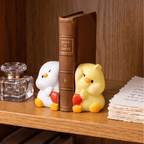 Two cute cartoon duck bookends holding a brown faux-leather book upright on a wooden shelf. One duck is white with yellow accents and holds a red heart, and the other is yellow with pink cheeks and yellow accents, also holding a red heart.