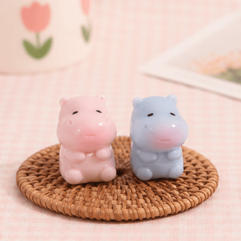 Two small hippo-shaped decorative figurines, one pink and one light blue, sitting side by side on a round woven coaster over a pale pink checkered surface.