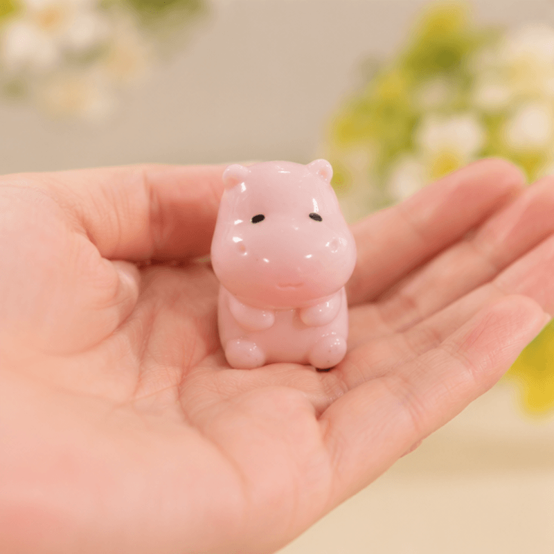 A small glossy pink hippo figurine resting on an open human hand, with a softly blurred indoor background and flowers.